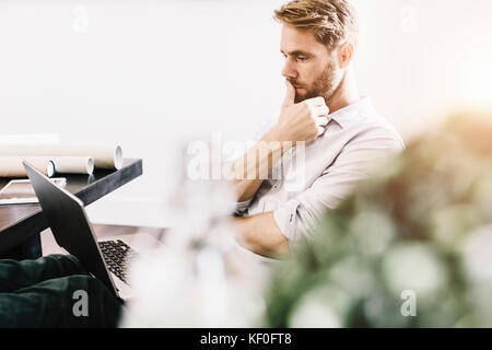 Portrait of pensive architect sitting with feet up at desk looking at laptop Stock Photo