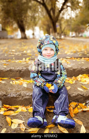 A child, a boy is sitting on the steps, a walk in the fall in the rain Stock Photo