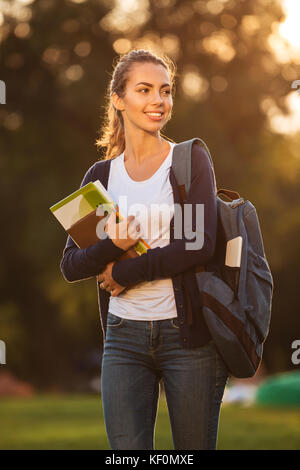 Portrait of a happy young schoolgirl with backpack holding books while walking and looking away outdoors Stock Photo