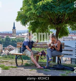 Couple sitting on a wooden bench overlooking the city of Cluj, the woman in playing a guitar and singing Stock Photo