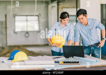 Young Asian engineers couple work together using notebook computer at building construction site. Civil engineering brainstorm meeting concept Stock Photo