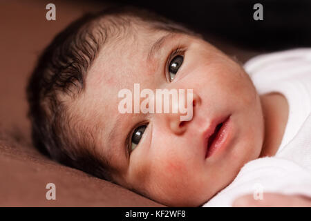 Close up portrait of a cute two weeks old newborn baby girl lying down, eyes open and looking around / newborn baby girl portrait lying down bed child Stock Photo