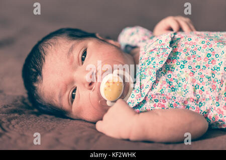 Portrait of a two weeks old newborn baby girl lying down with a pacifier or dummy, looking at camera with a floral dress / newborn baby pacifier dummy Stock Photo