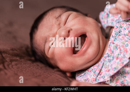Close up portrait of an unhappy cute two weeks old newborn baby girl lying down and crying hard, with cramps or colic, bellyache, stomach pain colics Stock Photo