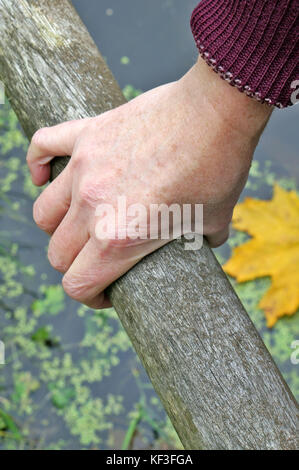 The elderly grandmother his right hand hold on to the railings of the wooden bridge across the village pond.  Maple leaf and water as background. Outd Stock Photo