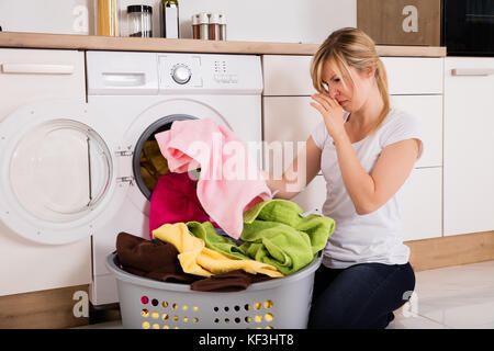 Young Woman Looking At Smelly Clothes Out Of Washing Machine In Kitchen Stock Photo