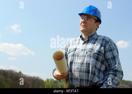 Construction site manager wearing blue helmet holding roll of building plans looking ahead shot over blue sky Stock Photo