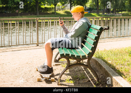 Boy Sitting On Bench With His Skateboard Using Mobile Phone Stock Photo