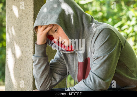 Unhappy Boy Wearing Grey Hoodie Sitting In Park Stock Photo