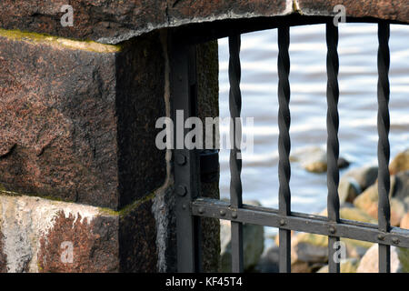 Close up of old prison bars and stone wall. Outside of the cell is stony beach and a sea. Stock Photo