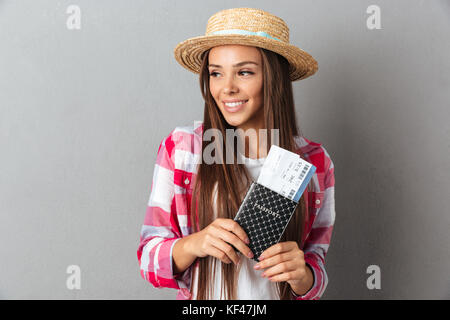 Close up portrait of a smiling happy woman traveller in straw hat holding passport with plane tickets looking away, over gray background Stock Photo