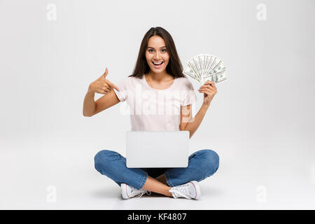 Portrait of an excited satisfied asian girl holding money banknotes while sitting with laptop computer on her lap and pointing finger isolated over wh Stock Photo