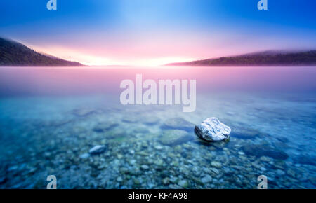 Long exposure of rock in lake at pebble beach with purple sunrise light. Stock Photo