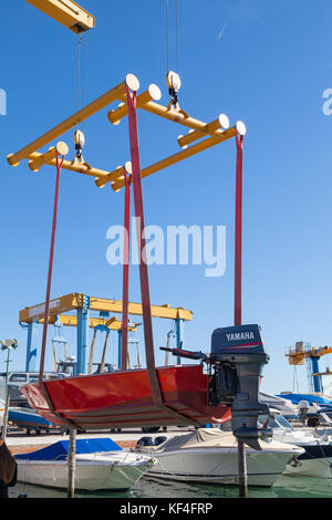 Small red boat with outboard motor suspended from a boat lift in a sling over water  at a marina during launch Stock Photo