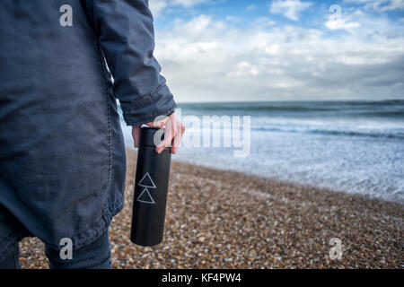 close up of a person holding a metal thermos flask while walking on a shingle beach Stock Photo