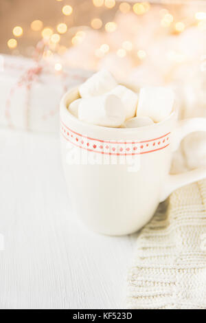 White Mug with Hot Chocolate Cocoa Drink and Marshmallows on Top. Sparkling Garland Lights in Background. Knitted Sweater Gift Box. Soft Pastel Colors Stock Photo