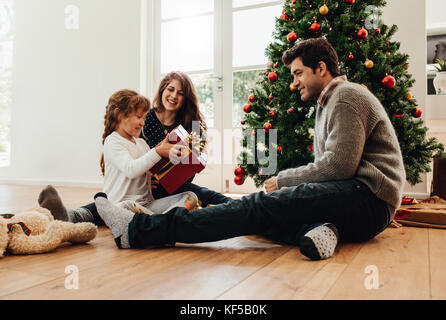 Parents presenting gift to their daughter sitting beside a Christmas tree. Little girl looks happy holding her Christmas gift. Stock Photo
