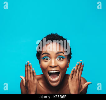 Excited young woman with vibrant eye shadow and nail paint. Female model showing her vivid makeup against blue background. Stock Photo