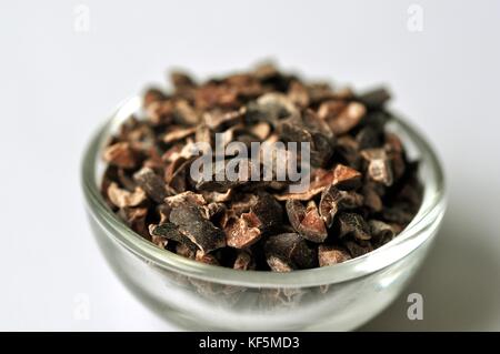 Raw cacao bean (Theobroma cacao) nibs in a glass bowl, isolated on white. Nutritious dietary supplement rich in minerals, vitamins, and fiber. Stock Photo