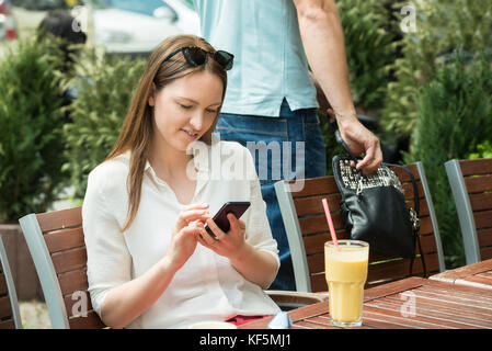 Male Thief Stealing Purse Of A Young Woman Using Mobile Phone Stock Photo