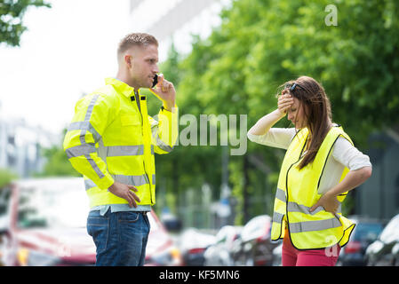 Stressed Young Couple Calling For Service On Mobile Phone With Broken Down Car On Street Stock Photo