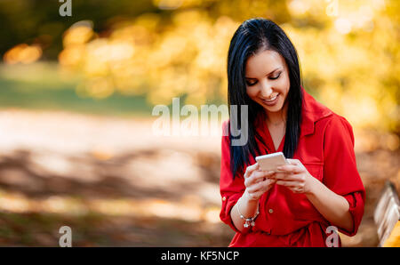 Happy young smiling woman reading message with pleasant news on smartphone in autumn nature. Stock Photo