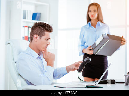 Young businessman holding phone and looking at laptop in the office. Businesswoman is standing behind him and holding binder. Selective focus. Focus o Stock Photo