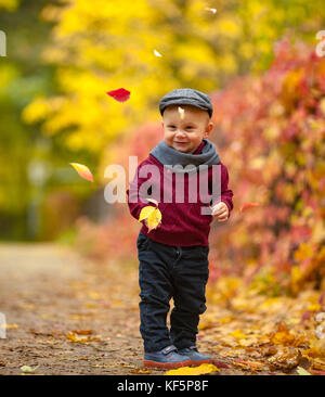 Little happy child boy wearing hat, scarf and sweater stands in park on background of flying colorful autumn leaves. Stock Photo