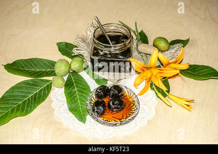 Glass jar with jam from walnuts on walnut leaves and orange lilies on a wooden table Stock Photo