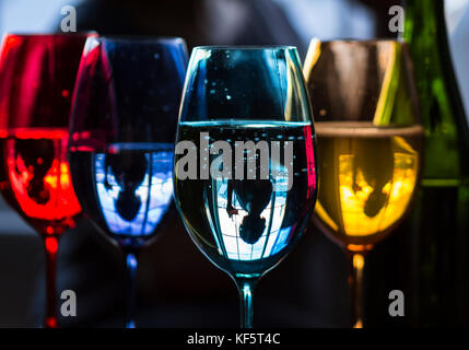 Portrait of woman having a drink by window. Upside-down reflection in colourful wine glasses. Stock Photo