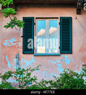 Tuscany, Italy. The wall of an old villa with flaking pink paint, and a window reflecting clouds in a blue summer sky Stock Photo