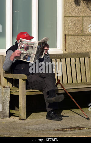 an elderly gentleman sitting on a bench reading a tabloid newspaper with his walking stick by his side in Saint Just, Cornwall, UK. Stock Photo