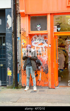 Man smoking and checking smart phone leaning against graffiti covered doorway in Brick Lane London England Stock Photo
