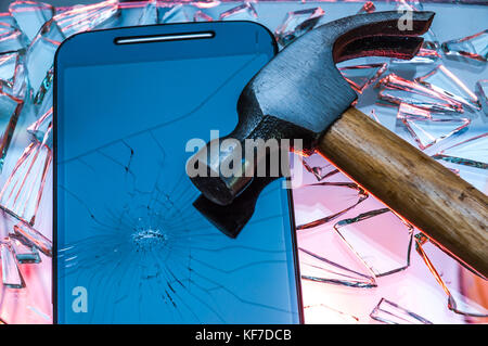Broken black smartphone with cracked screen lying on smashed broken glass with hammer on top of it Stock Photo
