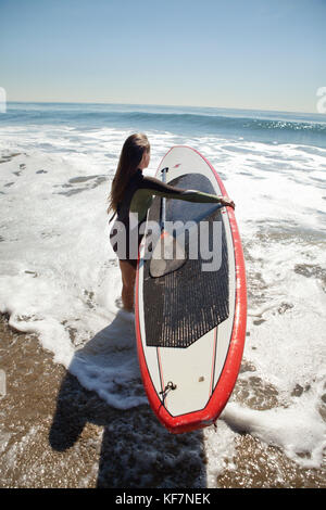 USA, California, Malibu, El Pescador Beach, an athletic woman carries her paddleboard into the Pacific Ocean Stock Photo