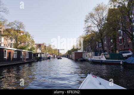 Point of view from a boat on a peaceful canal, flanked by moored houseboats and trees. Stock Photo