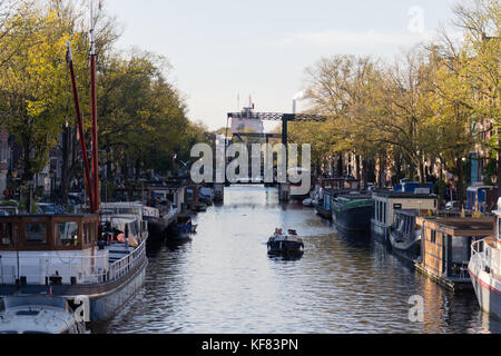 Boat sailing over Amsterdam canal with house boats on either side and bridges in the distance Stock Photo