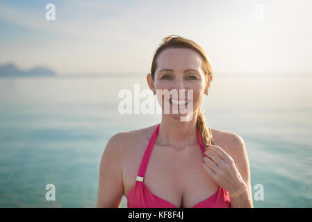 Happy smiling middle-aged woman holding on to her long hair standing with her back to the ocean and glow of the sun at the seaside as she enjoys a day Stock Photo
