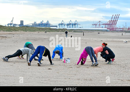 Instructor guiding people taking part in keep fit exercise class on Crosby beach Sefton Liverpool container port cranes beside River Mersea beyond UK Stock Photo