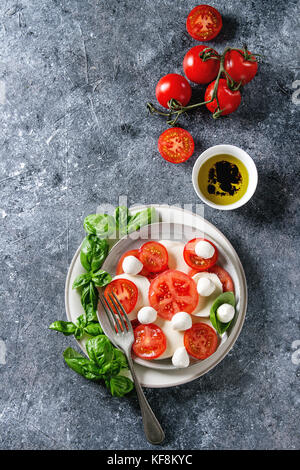 Italian caprese salad with sliced tomatoes, mozzarella cheese, basil, olive oil. Served in ceramic plate with ingredients above over gray texture back Stock Photo