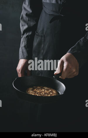 Man chef in black apron cooking pancakes in cast-iron pan. Holding pan in hand. Dark rustic style. Toned image Stock Photo