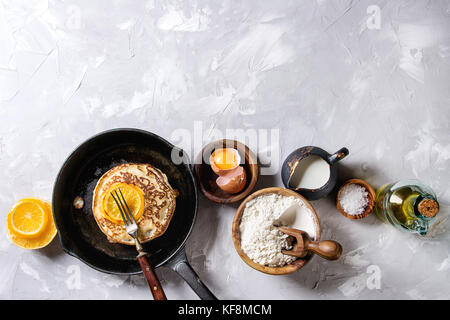 Homemade pancakes with fried orange in cast-iron pan and ingredients above. Wooden bowls of flour, yolk, salt, milk, olive oil over gray texture backg Stock Photo