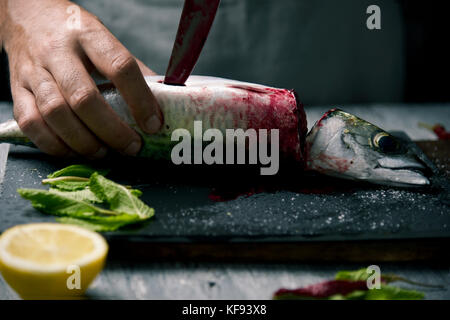 closeup of a young caucasian man cutting a raw fresh mackerel with a knife, on a slate tray placed on a rustic wooden table or countertop Stock Photo