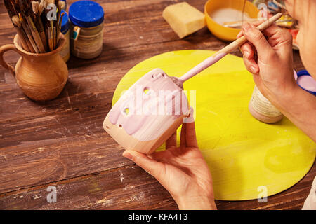 close-up artist and potter paints a clay bowl in gray color and a desktop on which lie kitschochki and other tools Stock Photo