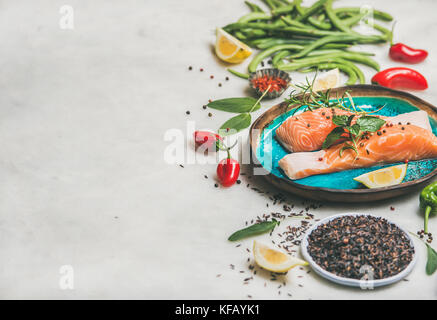Raw salmon fish fillet steaks with vegetables, greens, rice, spices and lemon in bright blue plate over grey marble background, copy space. Clean eati Stock Photo