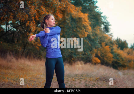 Slim caucasian jogger girl in blue shirt and black sports leggins performs warming-up  before jogging on the bright autumnal forest hill. Stock Photo