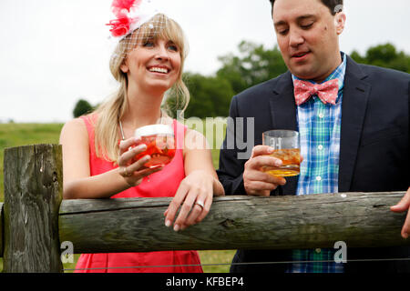 USA, Tennessee, Nashville, Iroquois Steeplechase, a young man and woman by a fence drinking Moonshine Cherry-Basil Blush and Tennessee Whiskey Stock Photo