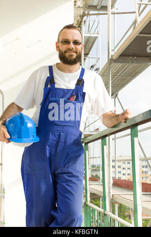Smiling construction worker stands with a protective helmet in hand against a background of scaffolding Stock Photo