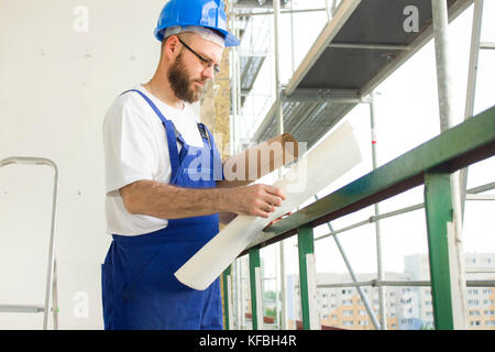 Construction worker in working outfit and in helmet stands at high altitude on construction site and inspects technical plans Stock Photo
