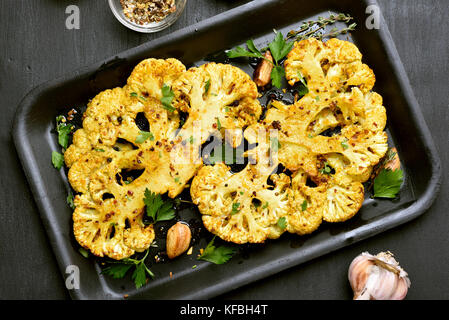 Baked cauliflower steaks with herbs and spices on baking sheet over black stone background Stock Photo
